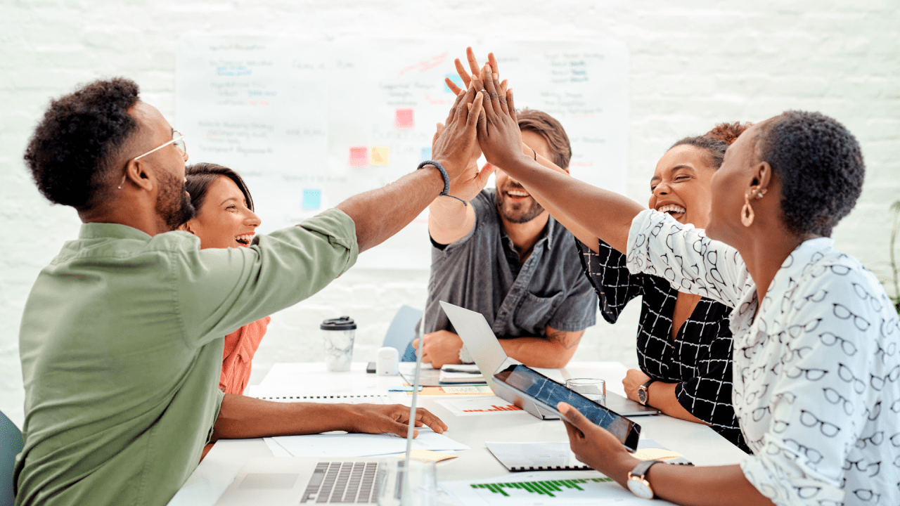 Grupo de personas sentadas alrededor de una mesa, sonriendo y chocando las manos en el centro