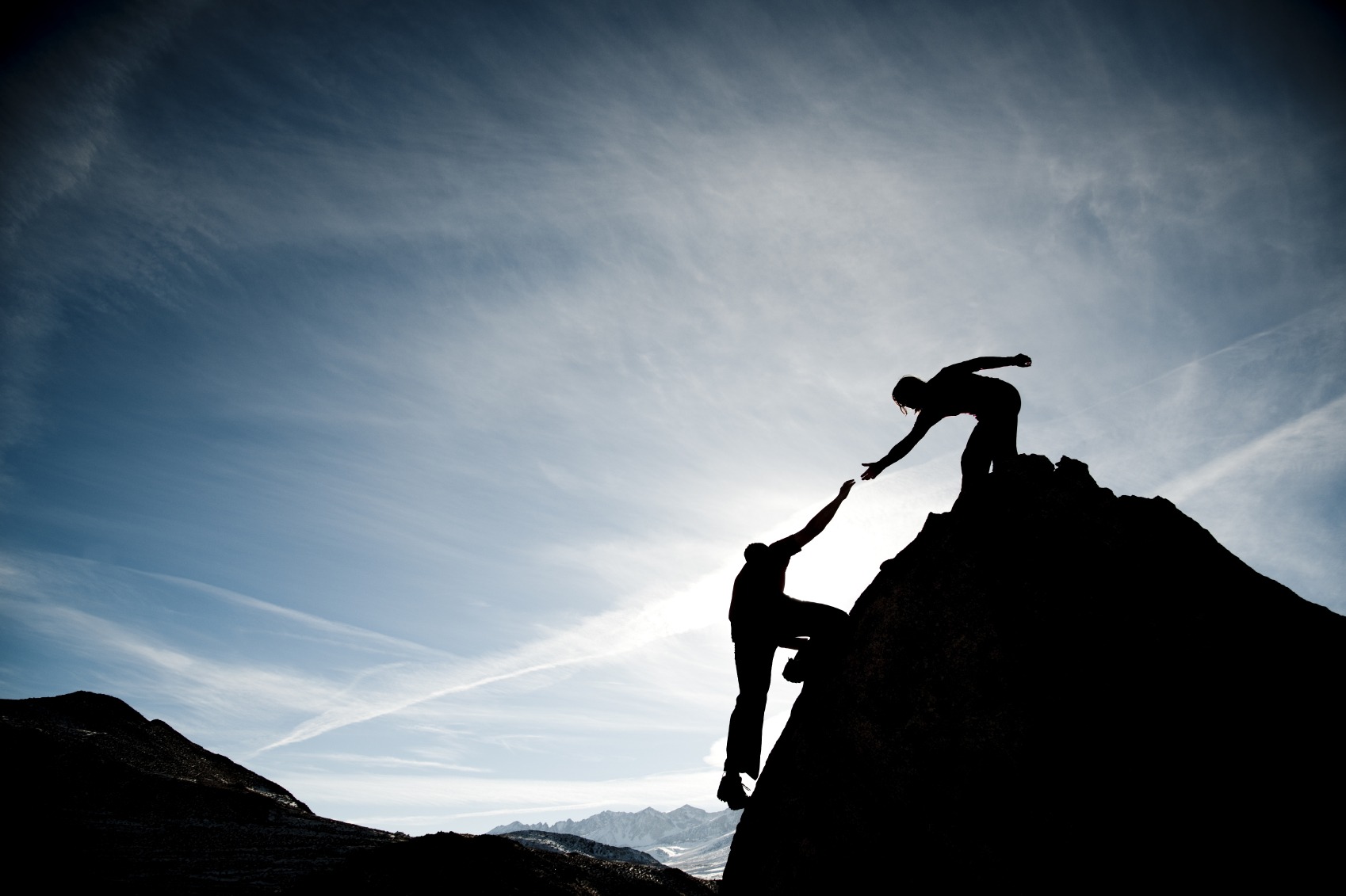 Una persona en el pico de una montaña dando la mano a otra que está intentando llegar a la cima